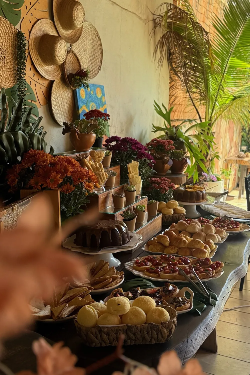 Mesa de evento montada com bolos, pães de queijo, tortinhas, flores e chapéus de palha na parede